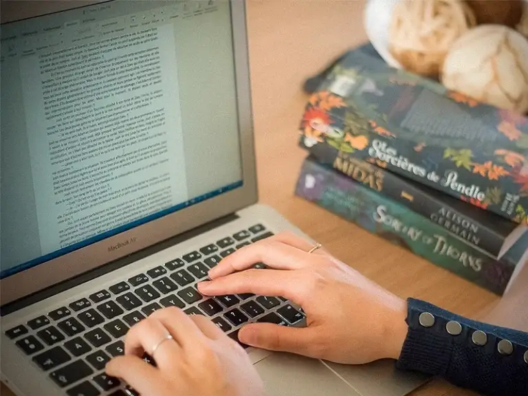 Autrice travaillant sur son manuscrit sur un MacBook Air, entourée de romans d’inspiration fantasy.